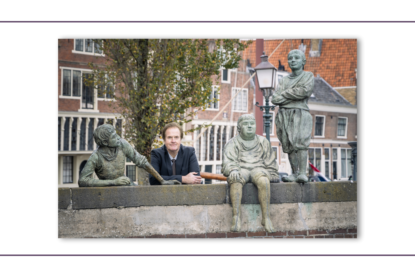 Een man in pak leunt op een stenen brug in de haven van Hoorn tussen drie bronzen beelden van de jongens van de Bontekoe. Op de achtergrond staan historische gebouwen, bomen en een lantaarn. De foto is genomen bij daglicht en heeft een rustige, klassieke sfeer.